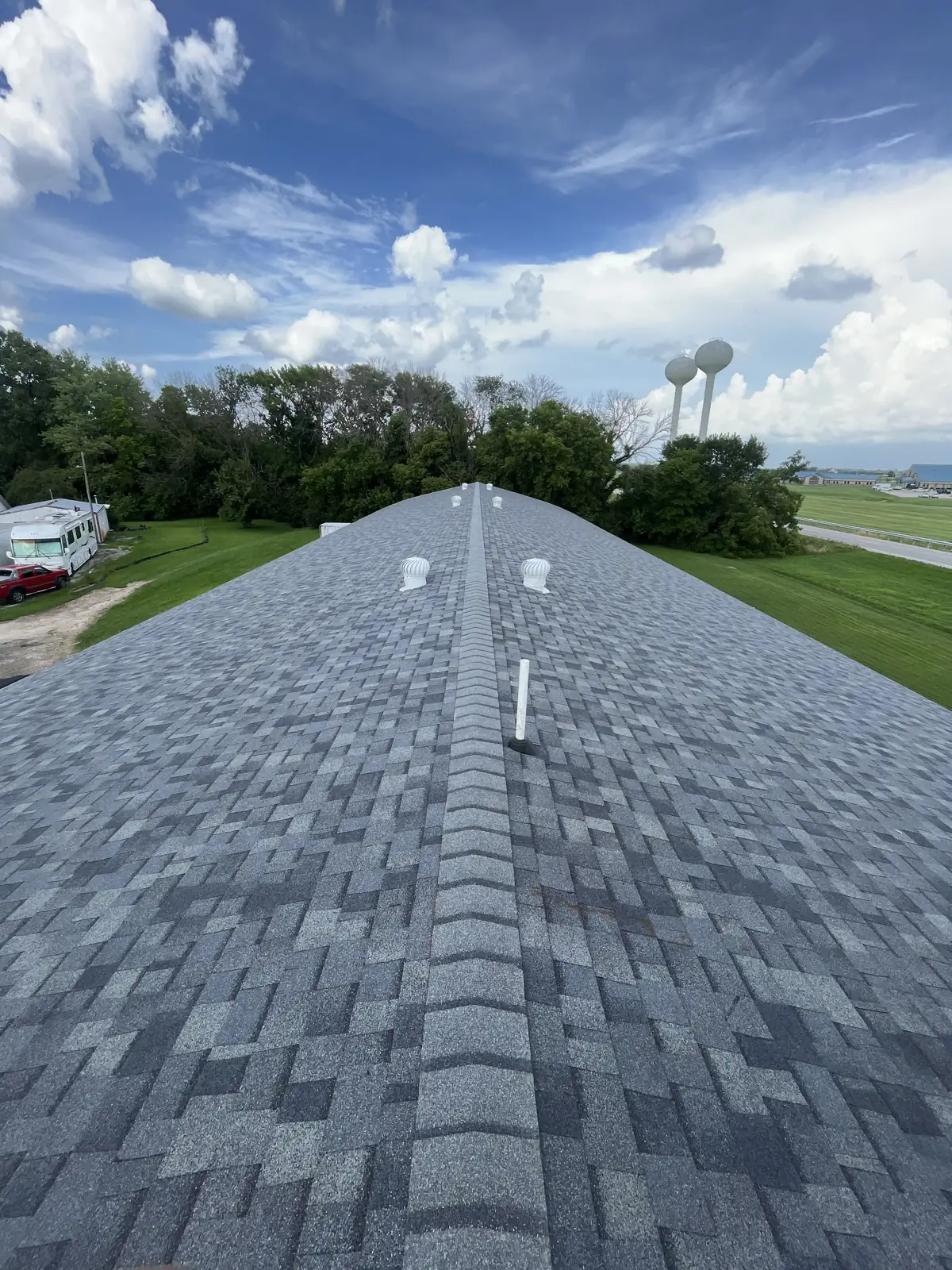 Finished gray architectural asphalt shingle roof with ridge cap and turbine vents on a Greenville, Illinois bowling alley, Greenville water towers in the background
