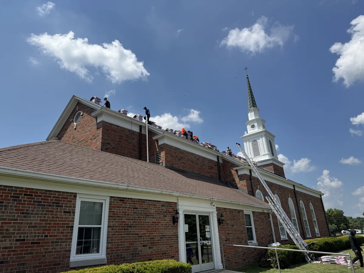 MPR Roofing crew installing new asphalt shingles on the roof of a brick Lutheran church with a white steeple in Greenville, Illinois